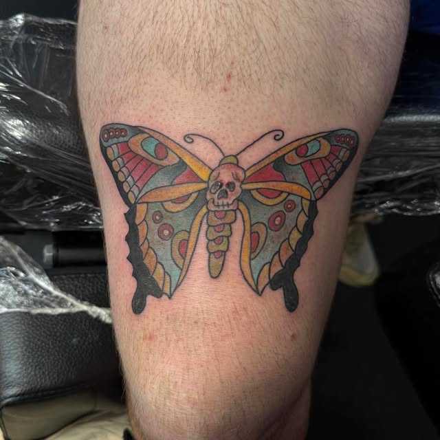 A traditional-style colour tattoo of a moth with a human skull on its body, wings patterned in red, teal and gold, on the outside of a lower leg.