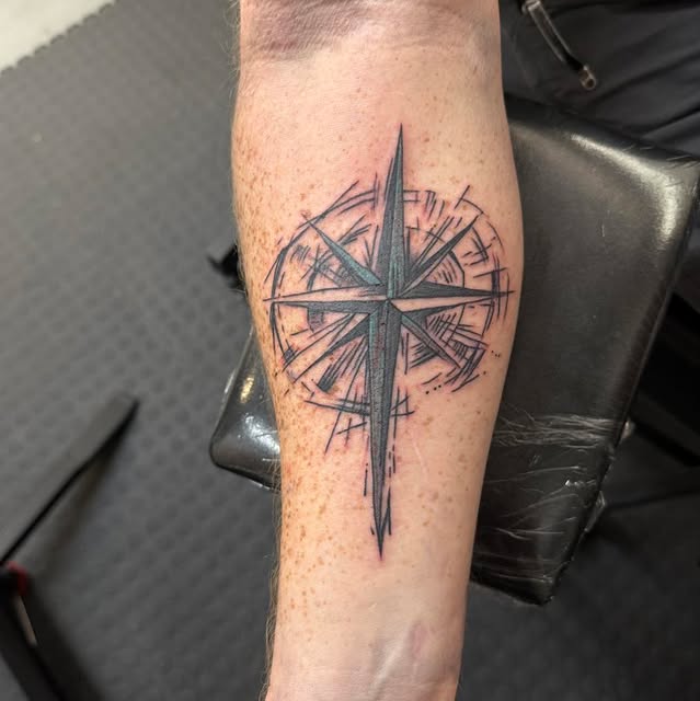 A black and grey compass rose on a forearm, drawn in a loose sketched line style with the long north point running down towards the wrist.