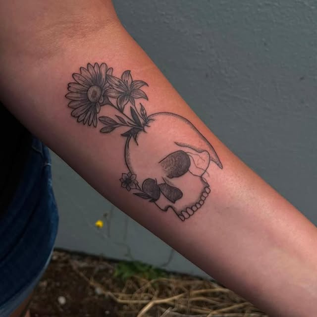 A fine-line black and grey tattoo of a skull with daisies and wildflowers growing across it, on the inside of a forearm, photographed outdoors.