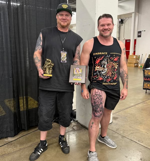 Two men standing side by side at a tattoo convention, one holding a small gold trophy and a framed award plaque, the other in a black vest, both heavily tattooed.