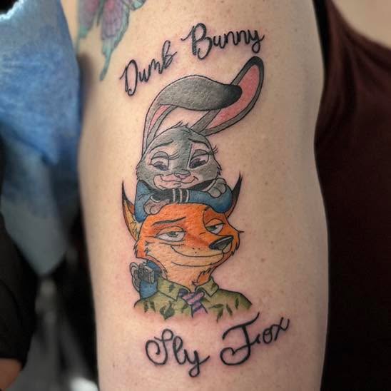 A colour cartoon tattoo of a rabbit above a fox on an upper arm, with the words Dumb Bunny arched above them and Sly Fox in script below.