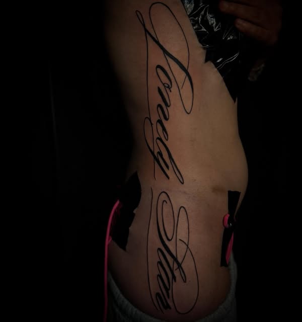 Large flowing script lettering tattooed down a ribcage, photographed against a dark background so the linework stands out.