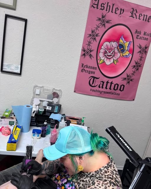 Ashley working at her station, in a pale blue cap with green hair, tattooing a client's arm. Her own shop banner hangs on the wall behind her, reading Ashley Renee, Ink Rich Tattoo, Lebanon Oregon.